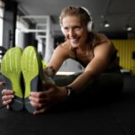 Eine Frau im Fitnessstudio dehnt sich mit Kopfhörern. Lächelnd sitzt sie auf dem Boden und berührt ihre neongrünen Schuhsohlen. Im Hintergrund sind verschiedene Geräte zu sehen.