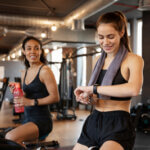 Zwei Frauen in Sportkleidung sitzen auf Heimtrainern in einem Fitnessstudio. Eine Frau blickt mit einem Handtuch um den Hals auf ihre Smartwatch, die andere hält eine Wasserflasche und lächelt. Im Hintergrund sind Fitnessgeräte zu sehen.
