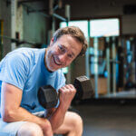 Mann in blauem T-Shirt und Shorts sitzt lächelnd in einem Fitnessstudio und hält in einer Hand eine Hantel. Im Hintergrund sind Fitnessgeräte und Spiegel zu sehen.