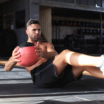 Ein Mann trainiert in einem Fitnessstudio und führt Russian Twists mit einem roten Medizinball aus. Er trägt schwarze Shorts und Turnschuhe, sitzt auf der Matte, die Beine leicht erhöht und den Oberkörper nach hinten gelehnt, um das Gleichgewicht zu halten.
