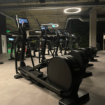 Eine Reihe moderner Crosstrainer in einem schwach beleuchteten Fitnessstudio mit Betondecke. Jedes Gerät verfügt über eine Tablet-Halterung. Kreisförmige Deckenleuchten sorgen für sanftes Licht. Im Hintergrund ist eine Wand mit hängenden Pflanzen zu sehen.