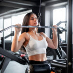 Eine Frau in weißem Sport-BH und schwarzen Leggings trainiert an einem Latzuggerät im Fitnessstudio. Konzentriert zieht sie die Stange zur Brust herunter. Das Fitnessstudio ist gut beleuchtet und hat große Fenster im Hintergrund.