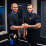 Zwei Männer in schwarz-blauen Fitnessanzügen lächeln und stoßen sich in einem Fitnessstudio an. Die Anzüge sind mit Kabeln ausgestattet, vermutlich für das EMS-Training. Im Hintergrund sind Fitnessgeräte und ein großes Glasfenster zu sehen.