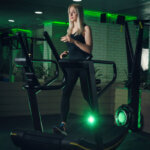 Eine Frau trainiert auf einem modernen Laufband in einem schwach beleuchteten Fitnessstudio mit grüner Beleuchtung. Sie trägt ein schwarzes Tanktop und Leggings. Der Raum hat verspiegelte Wände, die die Trainingsgeräte und die Atmosphäre reflektieren.