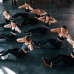 Eine Gruppe von Frauen in schwarzer Trainingskleidung führt in einem Fitnessstudio Bauchmuskelübungen im Sitzen durch. Sie sitzen in zwei Reihen auf einer großen, dunklen Trainingsmatte in gut beleuchteter Umgebung.