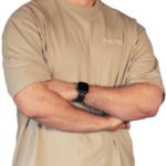 Eine Person mit Bart steht lächelnd mit verschränkten Armen da und trägt ein beiges T-Shirt und eine beige Hose. Am Handgelenk trägt sie eine Uhr. Der Hintergrund ist transparent.