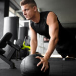 Ein Mann in einem schwarzen ärmellosen Hemd führt in einem Fitnessstudio eine Plankenübung mit den Händen auf einem Medizinball durch. Im Hintergrund sind Fitnessgeräte zu sehen.