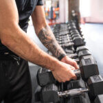 Ein Mann in schwarzem Shirt und Shorts sucht sich im Fitnessstudio eine Hantel aus einem Ständer. Sein rechter Arm ist detailreich tätowiert, Sonnenlicht fällt durch ein Fenster im Hintergrund in den Raum.