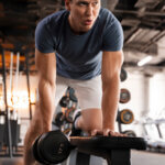 Ein Mann in blauem T-Shirt und weißen Shorts führt in einem Fitnessstudio eine Kurzhantelruderübung aus. Er stützt sich mit einer Hand auf eine Bank und zieht mit der anderen eine Hantel. Im Hintergrund sind verschiedene Gewichte und Fitnessgeräte zu sehen.