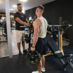 Zwei Männer im Fitnessstudio; einer trägt ein schwarzes T-Shirt und Shorts und hilft dem anderen, der ein beiges Tanktop und schwarze Shorts trägt, beim Gewichtheben. Sie stehen neben einer Bankpresse mit grün-schwarzen Gewichtsscheiben.