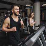 Ein Mann und eine Frau lächeln beim Joggen auf Laufbändern in einem Fitnessstudio. Beide tragen schwarze Trainingskleidung. Das Fitnessstudio ist mit verschiedenen Trainingsgeräten ausgestattet und großzügig und modern gestaltet.